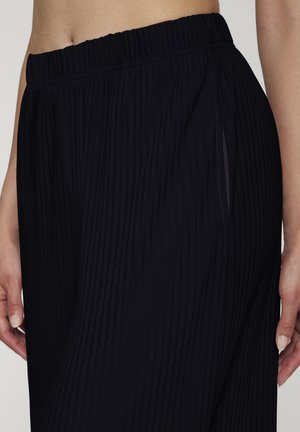 Close-up of a person wearing black pleated pants with an elastic waistband and side pocket, showing part of their midriff and hands.