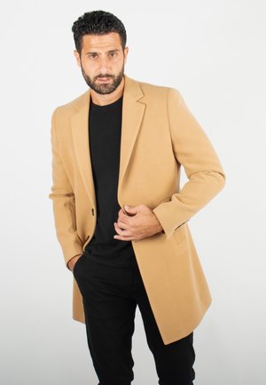 Manteau en laine couleur chameau avec un design ajusté, des revers en pointe et des poches avant, porté sur une chemise noire et un pantalon noir.