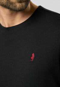 Schwarzes Baumwoll-T-Shirt mit Rundhalsausschnitt und einem kleinen roten gestickten Design einer Figur auf der linken Brustseite.