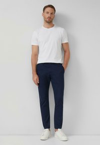 Muž stojaci proti bielej stene, oblečený v jednoduchom bielom tričku, námorníckych modrých nohaviciach a bielych teniskách, s jednou rukou v vrecku.