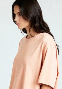 Per didelė, trumpomis rankovėmis marškinėliai iš minkštos persikų spalvos medvilnės. Turi apvalų kaklą ir nukritusias pečių siūles. Lygi tekstūra, minimalūs siuvimo detalės.