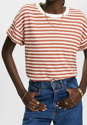 Personne portant une chemise à manches courtes blanche et orange rayée, un jean bleu taille haute, un collier en chaîne dorée, des bagues, un bracelet et des bracelets.
