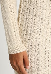 Svetr s kabelovým vzorem v krémové barvě, který má vyvýšené texturované vzory. Dlouhé rukávy s žebrovanými manžetami, ukazující měkkou pletenou látku.