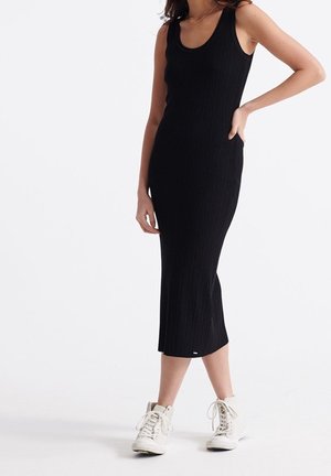 Femme portant une robe midi noire côtelée sans manches et des baskets blanches montantes, debout avec une main sur la hanche.