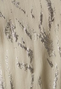 Beige Stoff mit strukturierter Oberfläche, der unregelmäßige silberne Metallmuster aufweist. Das Design erzeugt einen subtilen Glanz im gesamten Material.