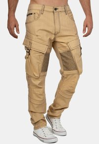 Pantalons cargo beiges avec plusieurs poches, accents texturés côtelés sur les cuisses et surpiqûres noires contrastantes. Matériel en mélange de coton.