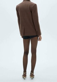 Ruskea houndstooth-kuvioitu bleiseri rakenteellisilla harteilla, yhdistetty mustiin shortseihin, läpikuultaviin ruskeisiin sukkahousuihin ja beigen värisiin korkeakorkoisiin sandaaleihin.