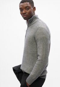 Jersey gris con textura y cuello con cremallera, confeccionado en tejido de punto texturizado. Cuenta con mangas largas y un diseño ajustado, combinado con pantalones negros.