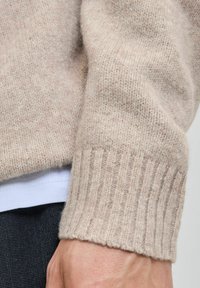 Lys beige striktrøje med ribbet manchet. Blød tekstur, let fuzzy finish, båret over en hvid skjorte, der afslører lagdelt stof.