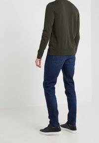 Olivově zelená mikina s dlouhými rukávy, žebrovanými manžetami a lemem, kombinovaná s tmavě modrými džíny s kapsami na zadní straně a zúženým střihem.