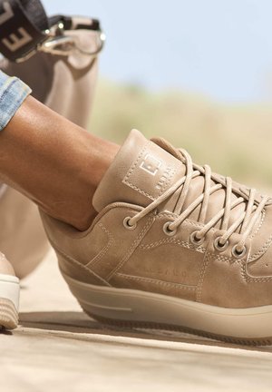 Beige leather sneaker with rounded toe, laces, and padded collar. Features a textured surface and subtle logo detail on the side.