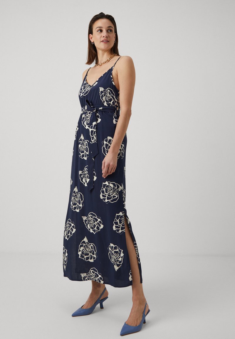 Robe longue marine à motifs floraux avec des roses crème, bretelles ajustables, fentes latérales et taille nouée. Portée avec des talons aiguilles bleus.