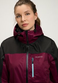 Burgundyröd och svart vattentät jacka med hög krage, justerbar huva och turkos dragkedjeficka. Har en logotyp på bröstet.