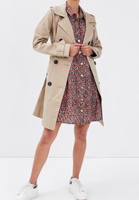 Woman wearing beige trench coat over red floral button-up dress, paired with white sneakers, standing against a plain white background.