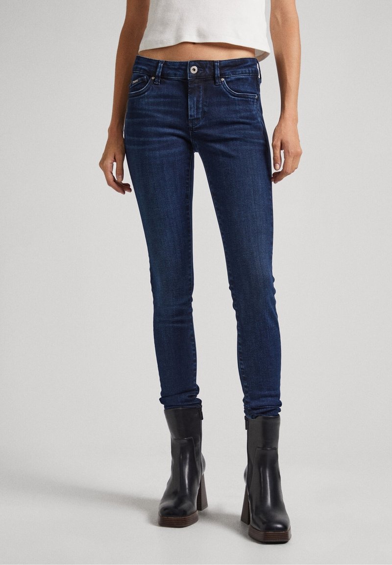 Skinny jeans in dark blue denim, featuring five pockets, belt loops, and a zip-fly front. Styled with black heeled ankle boots.