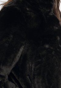 Sort faux fur jakke med en blød tekstur, høj halsudskæring og buede kanter, der fremhæver en glat, skinnende overflade.