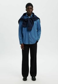 Ljusblå knapprad skjorta i denim, kombinerad med svarta vida byxor och en marinblå tröja hängande över axlarna. Svarta skor fulländar outfiten.