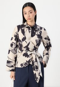 Camicia a motivi floreali in beige e nero, con colletto, maniche lunghe e dettaglio in vita annodato, realizzata in tessuto leggero.