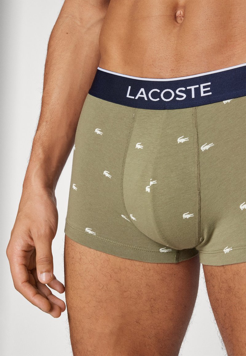 Oliwkowe bokserki z granatowym pasem w talii, ozdobione logo "LACOSTE", pokryte małymi białymi wzorami krokodyla i o miękkiej fakturze.