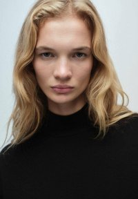 Crna dolčevita, mekane teksture, uskog kroja. Svijetlo plava kosa sa suptilnim valovima okružuje lice na neutralnoj pozadini.