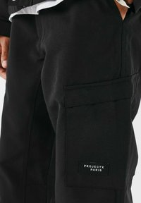 Pantaloni cargo neri con una tasca laterale e un'etichetta che recita "PROJECTX PARIS." Il tessuto appare robusto con una texture liscia.