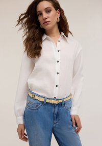 Camicia bianca con bottoni neri, maniche lunghe e texture semplice. Abbinata a jeans di denim azzurri e una cintura gialla.