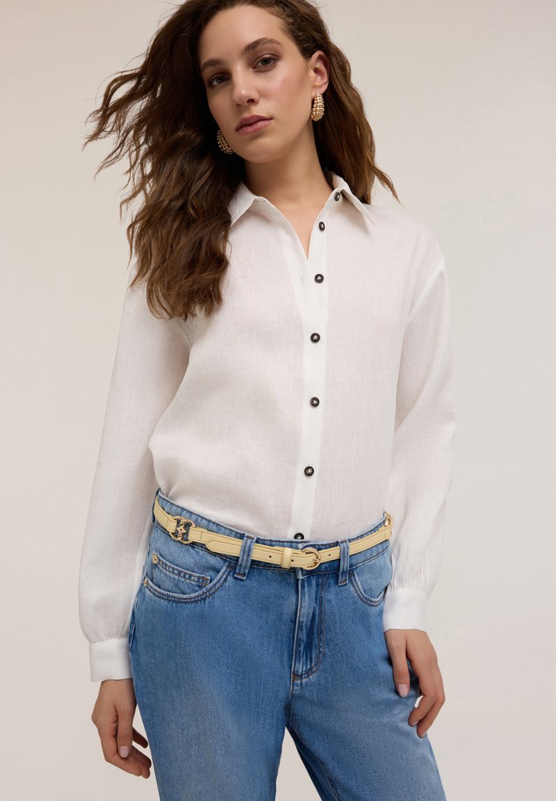 Camisa blanca de botón con botones negros, de manga larga y con textura lisa. Combinada con jeans de mezclilla azul claro y un cinturón amarillo.