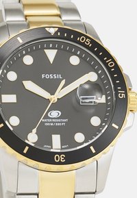 Montre Fossil avec un cadran noir, un bracelet en acier inoxydable doré et argenté, des marqueurs luminescents, une lunette rotative et un affichage de la date à 3 heures.