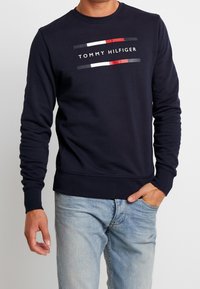 Felpa blu navy in cotone, con colletto rotondo, maniche lunghe e un logo "TOMMY HILFIGER" stampato con strisce colorate orizzontali.