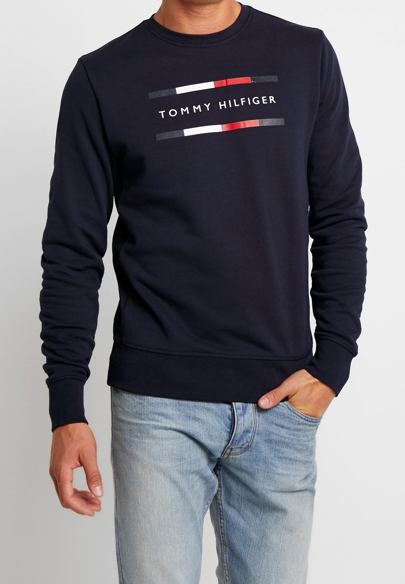 Felpa blu navy in cotone, con colletto rotondo, maniche lunghe e un logo "TOMMY HILFIGER" stampato con strisce colorate orizzontali.