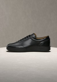 Μαύρο δερμάτινο χαμηλό παπούτσι τύπου sneaker με μαύρα κορδόνια και σόλα, τοποθετημένο πάνω σε ανοιχτογάλαζικη επιφάνεια με γκρι απλό φόντο.