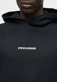 Gros plan sur une personne portant un sweat à capuche noir avec le mot "PEGADOR" imprimé en blanc sur la poitrine.
