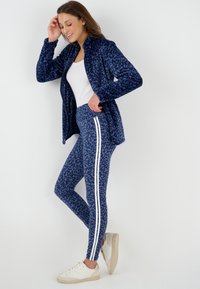 Donna con giacca leopardata blu navy e leggings abbinati con strisce bianche, top bianco e sneakers beige, in piedi su uno sfondo bianco.