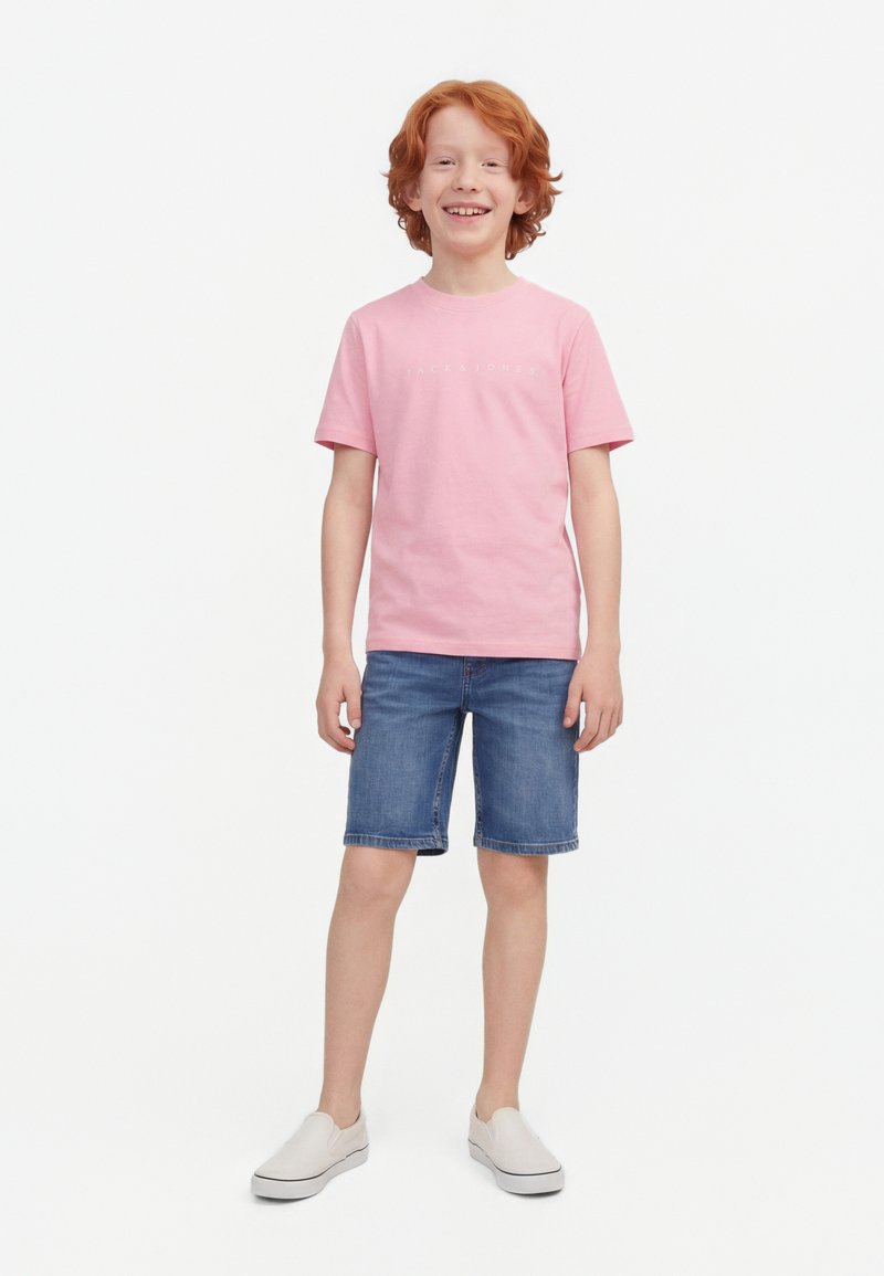 Garçon aux cheveux roux souriant, portant un T-shirt rose, un short en denim et des chaussures blanches sans lacets, debout devant un fond blanc uni.