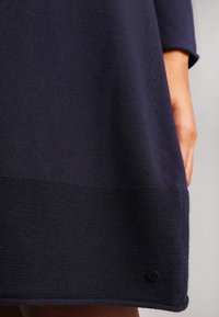 Abito lavorato a maglia blu navy con orlo texturizzato, corpo liscio e cuciture laterali in vista. Un piccolo logo si trova in basso a destra.