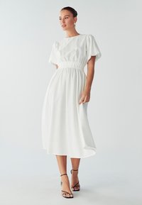 Kobieta w białej midi sukience z bufkami i zaznaczoną talią, w połączeniu z czarnymi sandałami na wysokim obcasie z paskami, stojąca na białej podłodze.