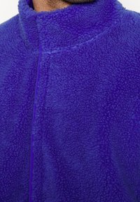 Polarowa kurtka w intensywnym purpurowym kolorze, z wysokim kołnierzem i miękkim, teksturowanym materiałem. Projekt zawiera minimalne detale szycia.