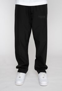 Black jogger pants made from lightweight fabric, featuring an elastic waistband with drawstring, side pockets, and a subtle logo patch.