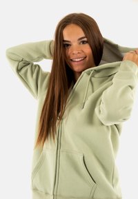 Sweatshirt à zip vert menthe en tissu doux, avec une capuche doublée, des cordons de serrage et des poches avant. Présente une texture lisse et une coupe décontractée.