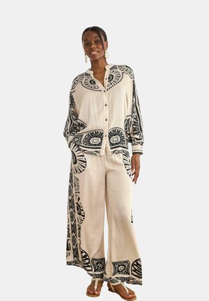 Mujer con una camisa crema de corte holgado abotonada y pantalones de pierna ancha con grandes estampados florales y geométricos negros, de pie y sonriendo.