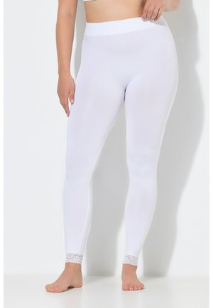 Donna con leggings bianchi a vita alta con bordatura in pizzo alle caviglie, in piedi a piedi nudi su uno sfondo semplice.