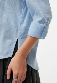 Chemise légère bleu clair à motif à carreaux, manches retroussées, fendue sur les hanches avec détail bouton aux poignets. Jupe grise visible en dessous.