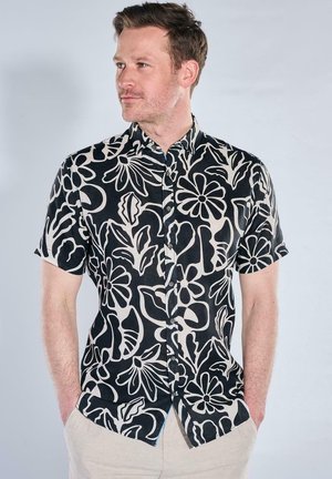 Mann mit kurzen braunen Haaren, der ein schwarzes kurzärmeliges Hemd mit großem weißem Blumenmuster trägt, Hände in den Taschen, schaut nach links.