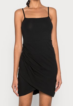 Kobieta w czarnej mini sukience na ramiączkach, z cienkimi paskami i asymetryczną marszczoną spódnicą na jednolitym tle.