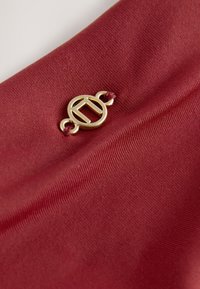 Emblème circulaire doré avec un motif géométrique fixé à un tissu rouge texturé par deux petits nœuds.