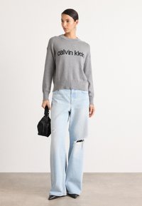 Hallhalline kootud sviiter, millel on "calvin klein" logo, koos helesiniste rebenenud laia säärega teksade ja musta tekstuuriga käekotiga. Teravad mustad kontsad.