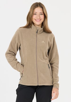 Jeune femme souriante portant une veste en polaire beige zippée, les mains dans les poches, sur un fond blanc uni.