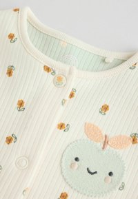 Lys cremefarvet ribbet stof beklædning med grønne og gule blomster mønstre, der har et broderet æble design med et smilende ansigt og blad detaljer.