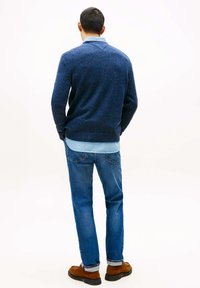 Marineblauer Strickpullover mit gesprenkelter Textur, über einem hellblauen Hemd getragen, kombiniert mit blauen Jeans und braunen Schuhen, von hinten betrachtet.