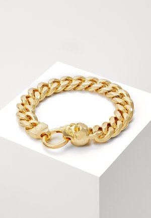 Bracelet à chaîne épaisse ton or avec un fermoir en forme de crâne reposant sur un bloc géométrique blanc sur un fond neutre.